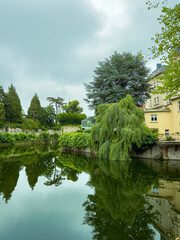 In a tranquil countryside, a pond reflects lush greenery and overcast skies, creating an idyllic...
