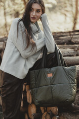 Woman in a Grey Turtleneck Sweater, Leaning on a Pile of Wood, Presenting a Large Green Quilted Bag