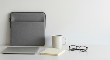 Minimalist workspace setup featuring a laptop, laptop sleeve, notebook, mug, and glasses against a white background.