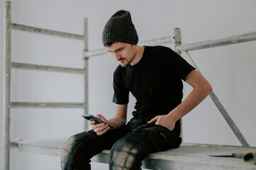 A young worker sits on scaffolding with a smartphone in his hands.