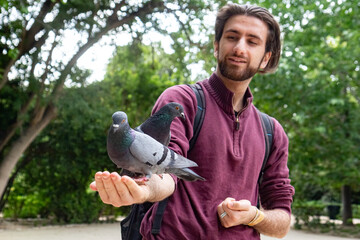 Young man smiling joyfully while holding multiple pigeons on his arm in a city park. Beautiful moment of harmony with nature.

