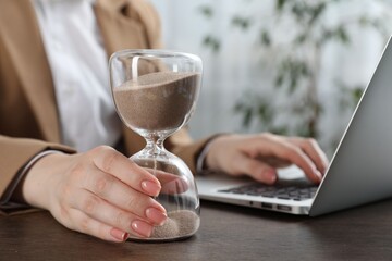Deadline. Woman with hourglass and laptop working at wooden table in office, closeup