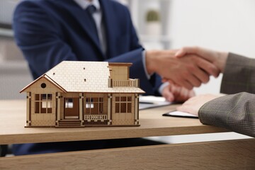 Real estate agent and client shaking hands at desk in office, focus on house model