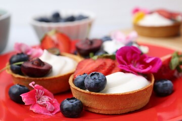 Sweet tartlets with berries and flowers on plate against light background, closeup. Delicious dessert