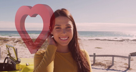 Smiling woman tucking hair on sandy shore by fence, with convertible and red heart graphic