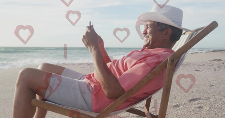 Scrolling smartphone senior man wearing straw hat sitting on beach chair, with floating heart icons