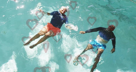 Floating senior couple wearing rash guards and trunks in backyard pool, with heart overlays