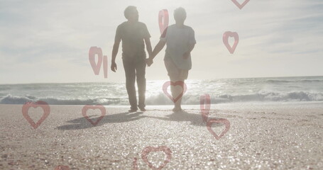 Holding hands senior couple walking along sunlit shoreline, with red heart and location-pin icons