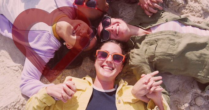Smiling group of four women lying in circle on sandy beach, with bright sunglasses, summer clothing