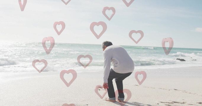 Bending senior Hispanic man picking up pink flamingo stake on beach, with floating heart overlay - Powered by Adobe