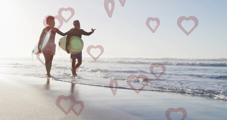 Walking couple in swimwear carrying surfboards with leashes along beach at low tide, heart graphics
