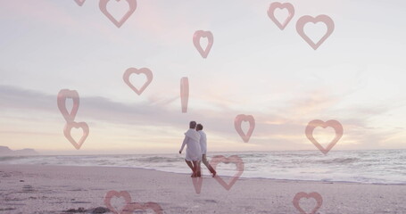 Walking senior couple wearing light clothes arm in arm on sandy beach at sunset, evening glow