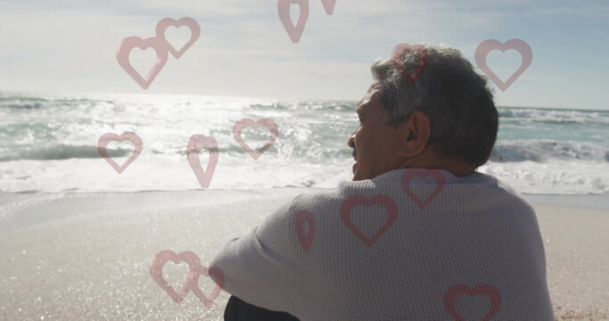 Sitting senior Hispanic man gazing at ocean waves on shoreline, with red heart overlays, copy space