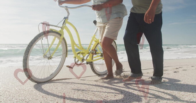 Strolling senior couple walking barefoot along sandy shoreline, with pastel yellow cruiser bicycle