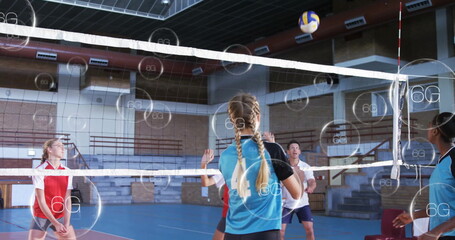 Teen volleyball player spiking ball over net in sports hall, wearing blue-black jersey, 6G overlays