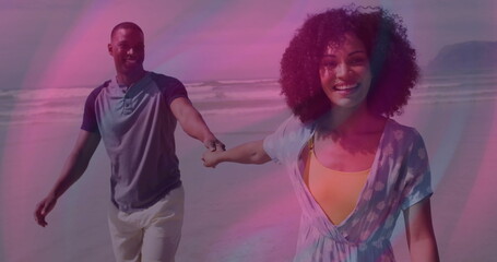 Walking couple holding hands along wet sand by ocean, wearing henley shirt and floral beach dress