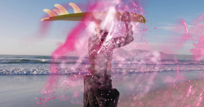 Walking surfer wearing black wetsuit holding surfboard overhead on beach, with swirling pink powder - Powered by Adobe