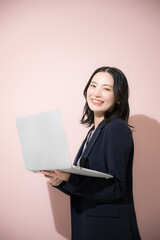 パソコン持ってスーツを着た立つカメラ目線の若いかわいい女性のビジネス上半身　ピンク背景の営業などリクルートのイメージ	縦