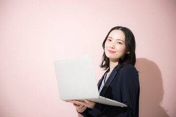 パソコン持ってスーツを着た立つカメラ目線の若いかわいい女性のビジネス上半身　ピンク背景の営業などリクルートのイメージ	