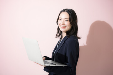 パソコン持ってスーツを着た立つカメラ目線の若いかわいい女性のビジネス上半身　ピンク背景の営業などリクルートのイメージ	