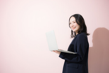 パソコン持ってスーツを着た立つカメラ目線の若いかわいい女性のビジネス上半身　ピンク背景の営業などリクルートのイメージ	
