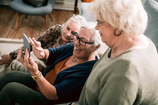 Senior women laughing together while looking at smartphone on living room couch