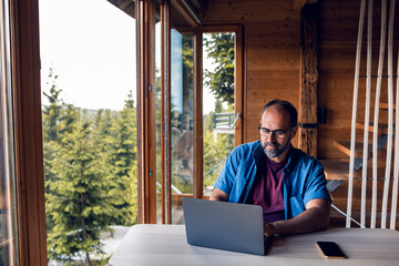 Man working remotely on laptop in forest cabin with scenic view