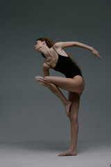 graceful woman dancer in black leotard performs contemporary dance pose with extended arms and leg against plain gray background in studio setting