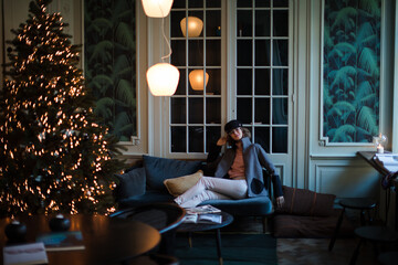 Fashionable woman sitting on cosy sofa next to lighted Christmas tree. in elegant environment....