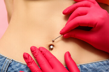 Professional doing piercing of young woman's navel on pink background, closeup
