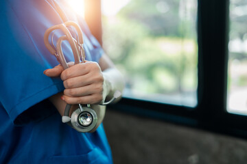 Close-up of a healthcare worker in blue scrubs holding a stethoscope, with sunlight streaming...