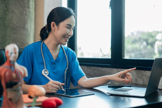 Healthcare professional in blue scrubs working on a laptop in a medical office, surrounded by anatomical models.