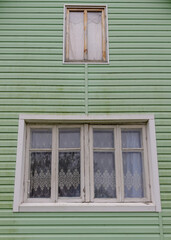 Close-up of two old windows with lace curtains on a pale green wooden house facade, showing vintage rural architectural style.
