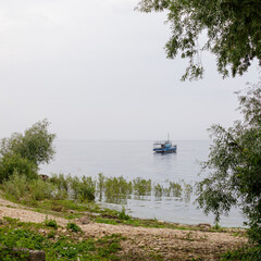 Small blue vessel calmly floats on the reflective surface of Lake Ilmen, viewed from a green and rocky shoreline with trees.