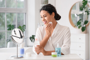 Beautiful young African-American woman removing makeup with cotton pad and looking at mirror in bathroom © Pixel-Shot