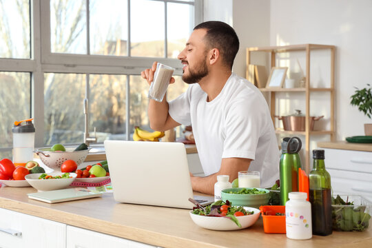 Sporty young man with fresh vegetables, vitamins and laptop drinking healthy smoothie in kitchen. Healthy food concept