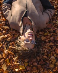 Head shot of beautiful woman laying up side down on natural forest carper made out of autumn leaves, while wearing glasses and fashionable fall clothing during natural sunlight and scenic coloured nat