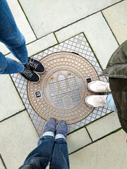 Top view of three pairs of shoes standing around a decorative manhole cover in Hamburg. Urban detail with people gathered in a circle on a paved street.