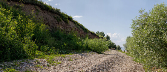 Pebbled path running along the base of an eroded grassy cliff, surrounded by lush greenery and...
