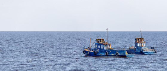 Pair of weathered blue fishing boats anchored in open water under a bright sky, captured from a...