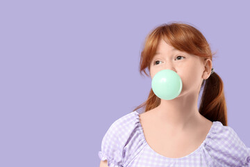 Cute ginger teenage girl blowing bubble gum on lilac background