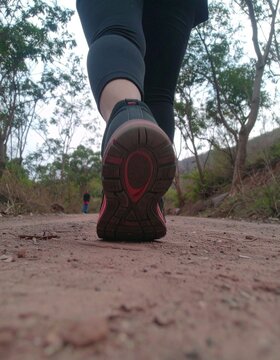 A POV (point-of-view) illustration from behind captures the lower half of a jogger in motion, focusing on their running shoes as they stride along a tree-lined park path. 