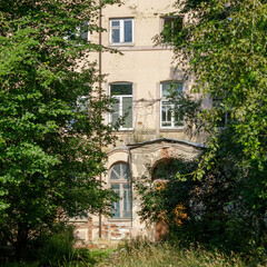 A decaying multi-story building partially obscured by dense greenery, showing cracked walls, broken...