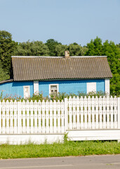 Traditional blue wooden cottage with a gray roof and overgrown garden, seen behind a white...