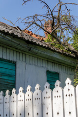 Weathered rural house with faded shutters and tiled roof stands behind a white decorative fence,...
