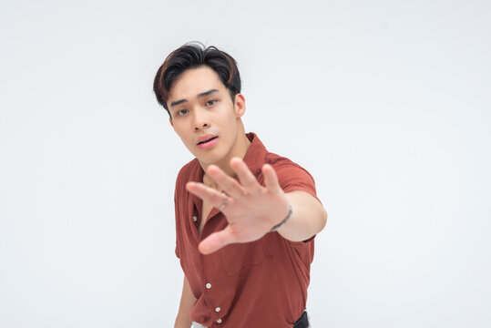 An unhappy young Asian man extending both hands forward in a ‘wait’ gesture, looking displeased. Isolated on a white background.