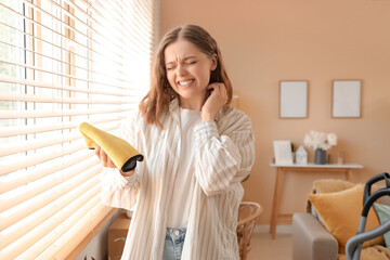 Young woman scratching herself while cleaning window blinds. Allergy concept
