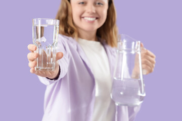Young woman with glass and jug of water on lilac background, closeup