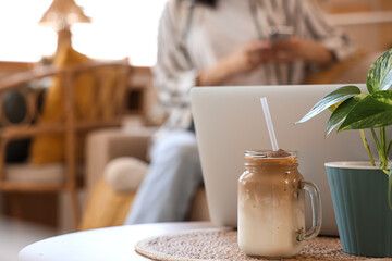 Glass jar of iced latte on table against woman using mobile phone at home, closeup