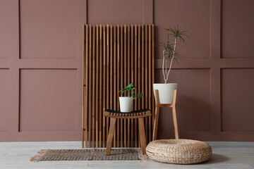 Wicker pouf, folding screen and houseplants near brown wall in interior of room, closeup